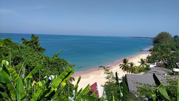 Ein Strand mit klarem, blauem Wasser, Palmen und ein paar Gebäuden im Hintergrund.