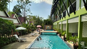Ein Swimmingpool mit Liegestühlen und Sonnenschirmen, umgeben von Grünpflanzen und einem Gebäude mit grüner Fassade.