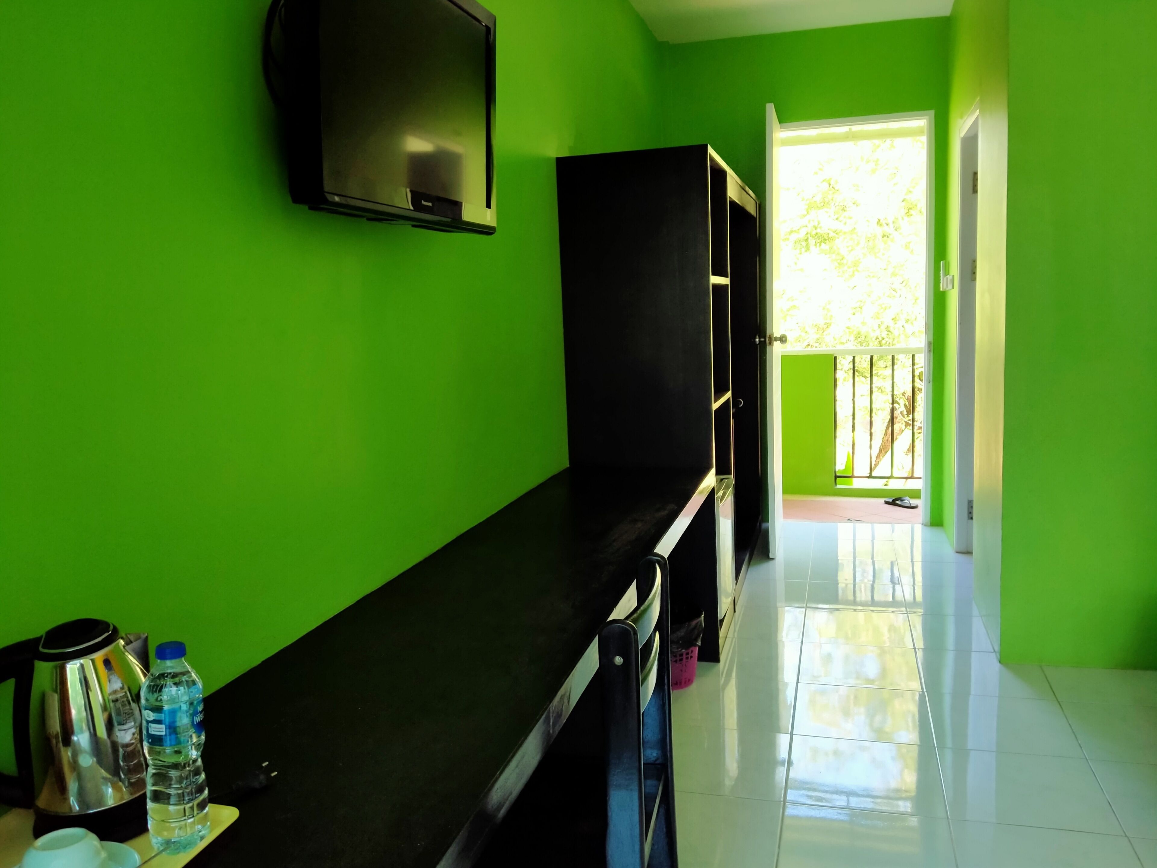 A room with a green wall, a flat-screen TV mounted on the wall, a black cabinet, a water bottle, and a kettle on a table.