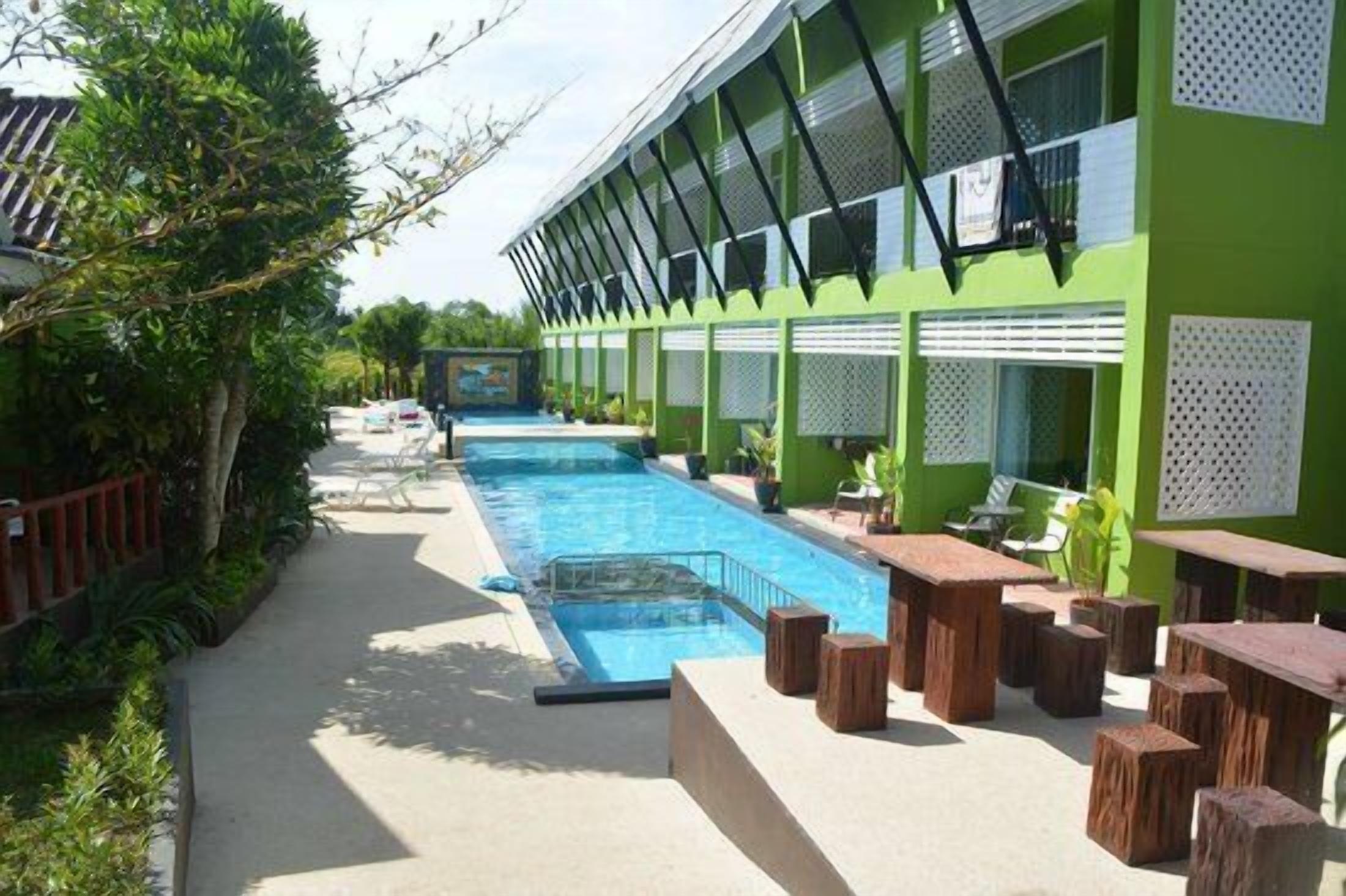 A poolside area with a long pool, wooden seating, and a green building with large windows.