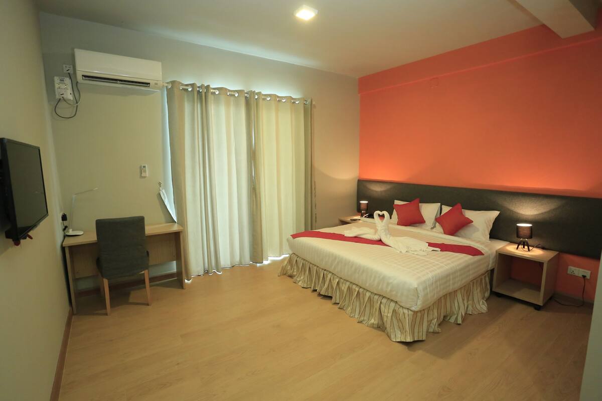 A bedroom with a bed, a desk, a chair, a TV, and a nightstand with a lamp.