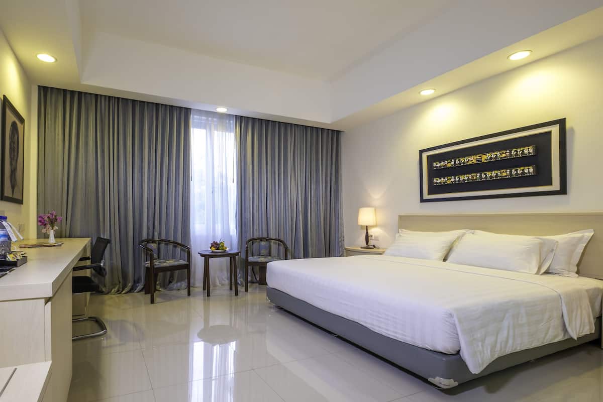A hotel room with a bed, a desk with a chair, a small table with a chair, a TV, and a window with curtains.