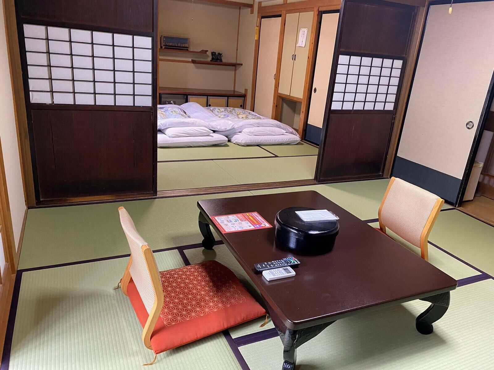 A traditional Japanese room with tatami flooring, a low table with a cushion, a bed with futons, and sliding doors.