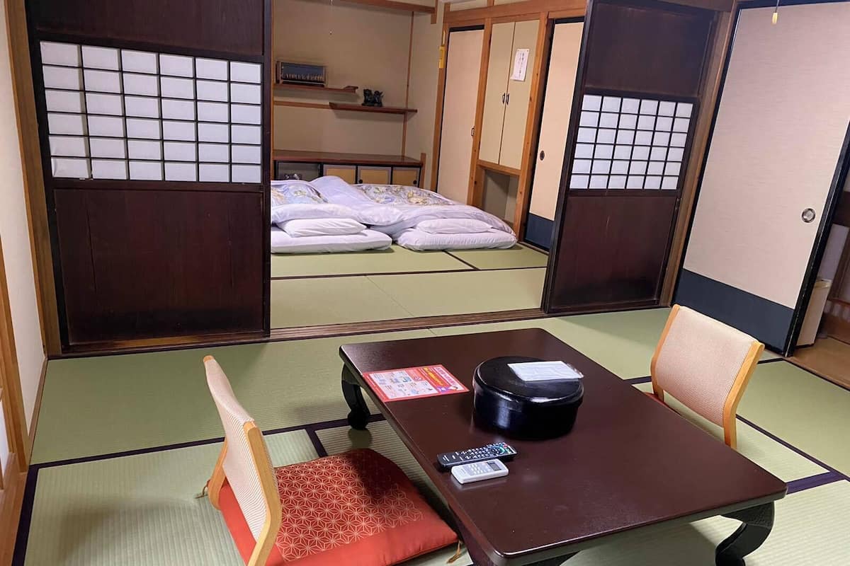 Una habitación japonesa tradicional con suelo de tatami, una mesa baja con cojín, una cama con futones y puertas correderas.