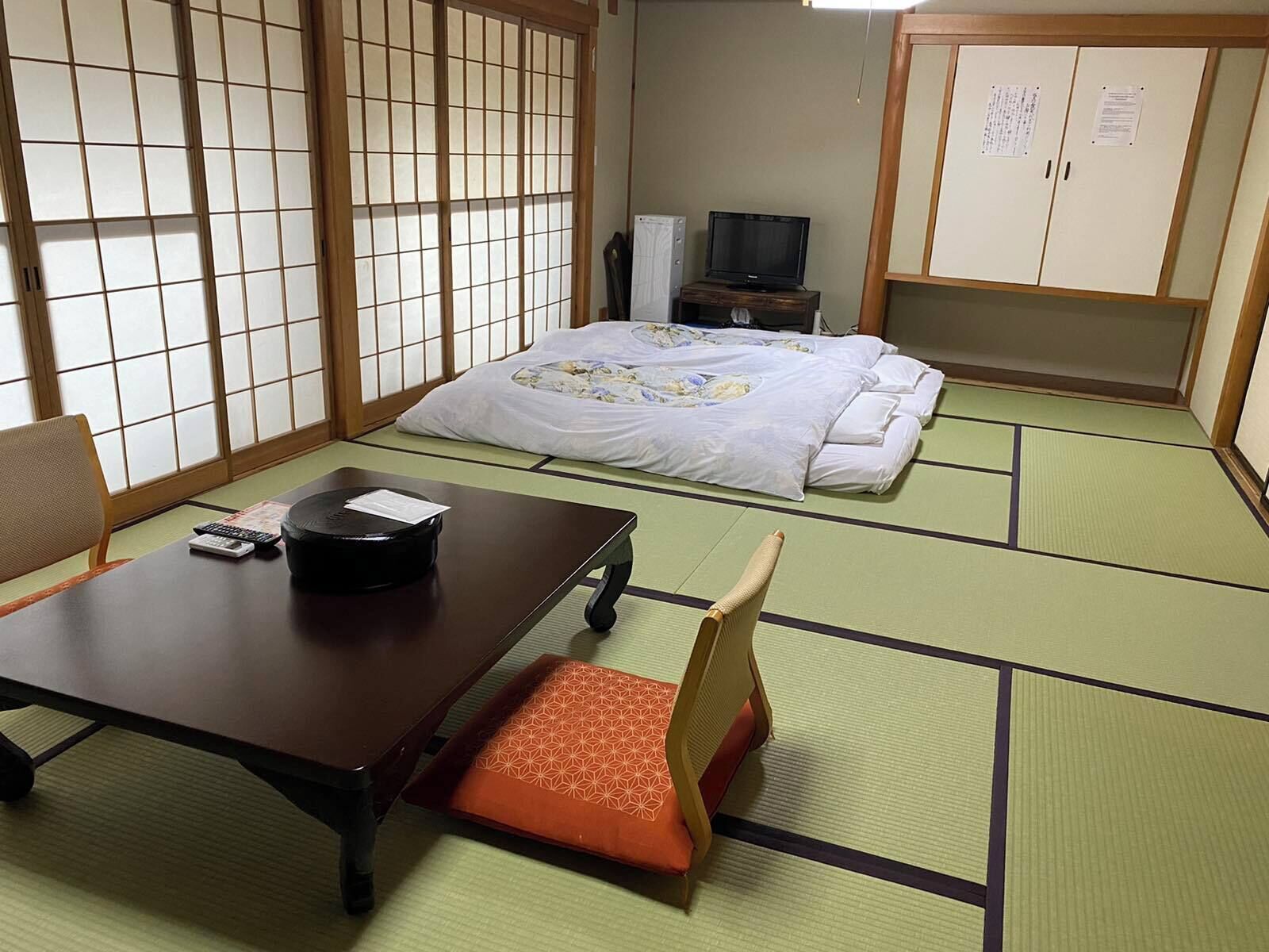 A traditional Japanese room with tatami flooring, a bed with a floral bedspread, a television on a wooden stand, and a desk with a remote control.