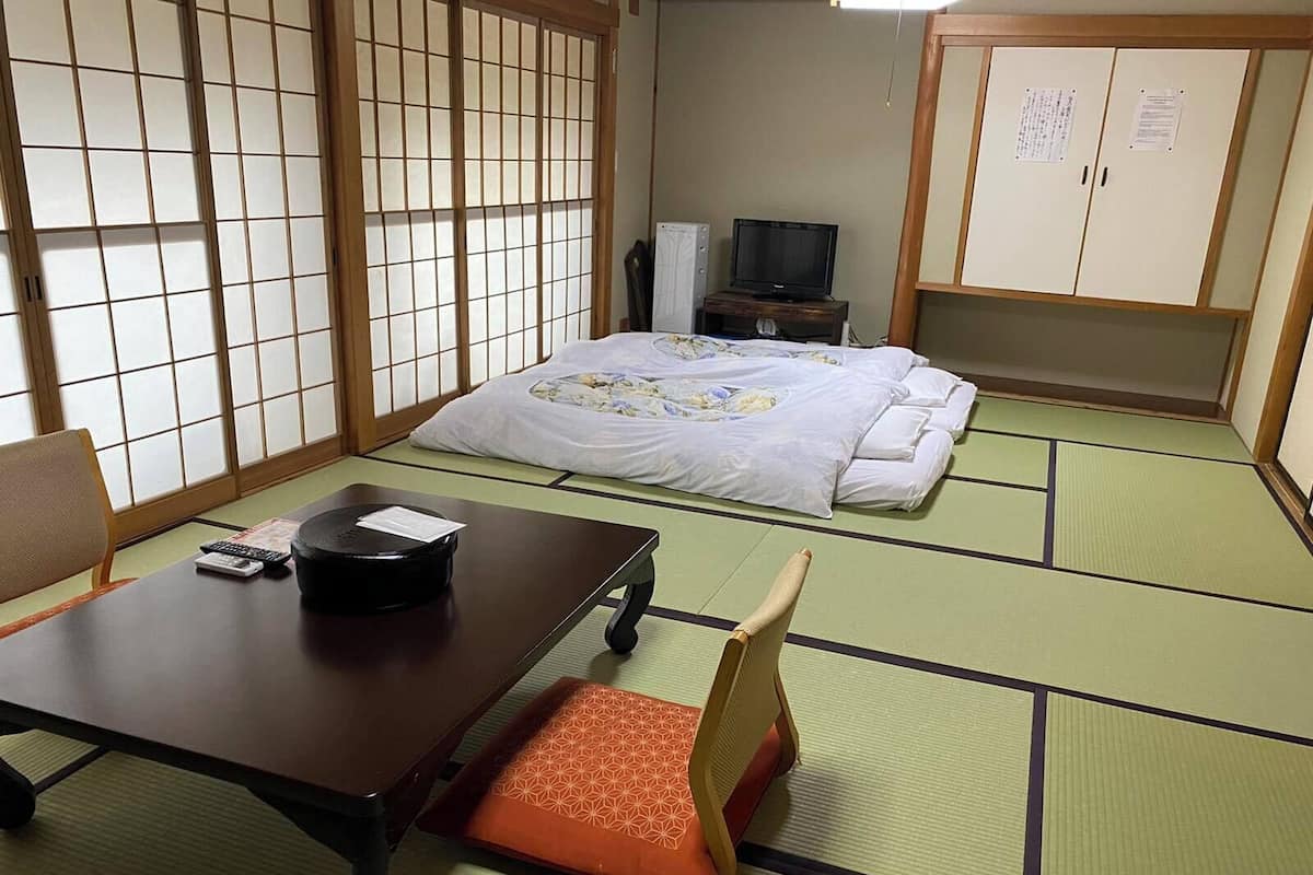 Una habitación japonesa tradicional con suelo de tatami, una cama con colcha floral, un televisor sobre un soporte de madera y un escritorio con un control remoto.