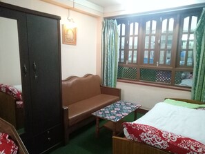 A room with a brown sofa, a small table with a floral tablecloth, a bed with a white and red patterned bedspread, a wardrobe, and a window with green curtains. - Swastik Guest House (Bhaktapur)