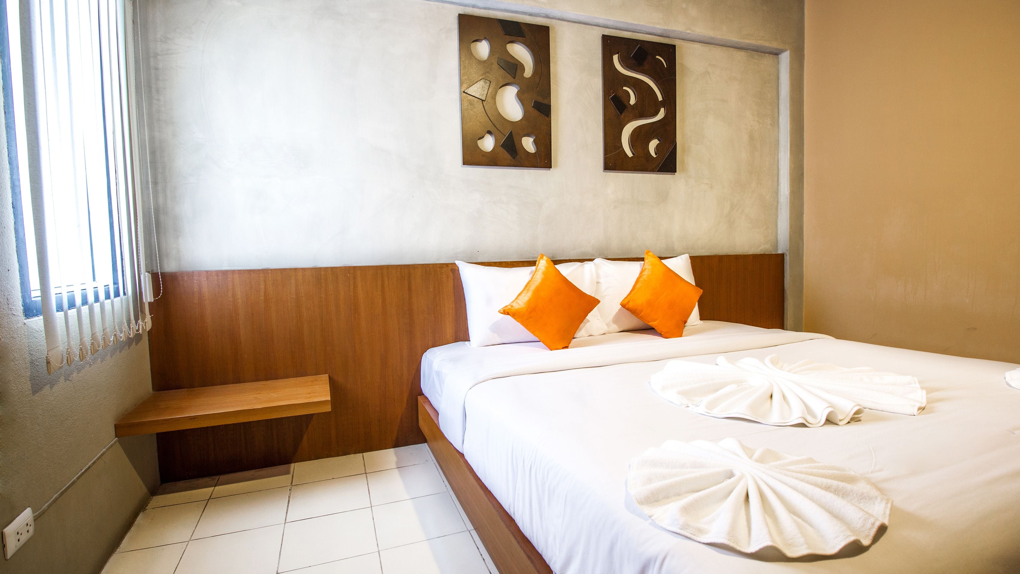 Habitación de hotel con cabecera de madera, dos almohadas naranjas, ropa de cama blanca y un perchero con toallas.