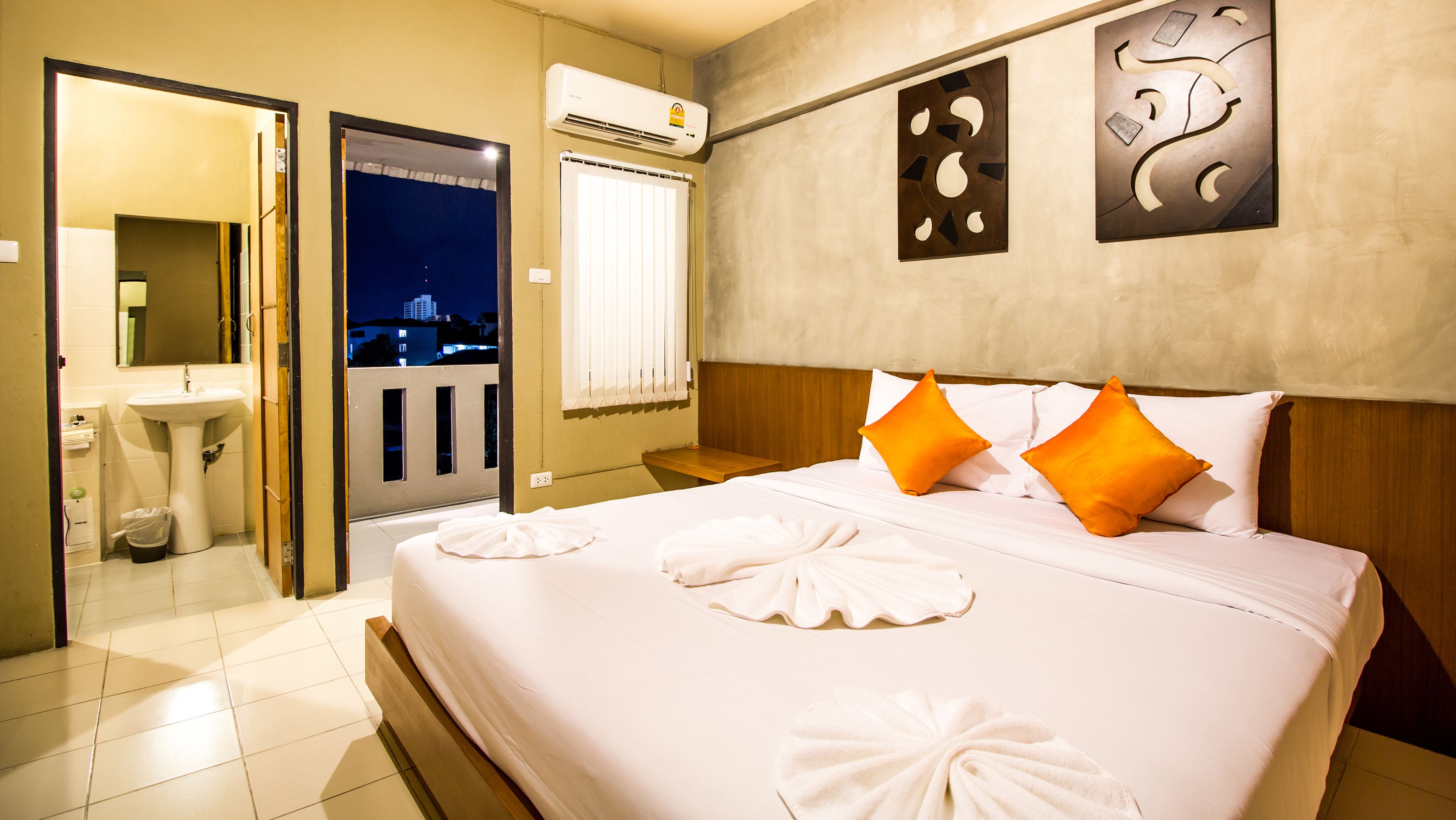 Habitación de hotel con cama, dos almohadas, mesita de noche, ventana con persianas, aire acondicionado instalado en la pared, baño con lavamanos y espejo, y vista nocturna de la ciudad.