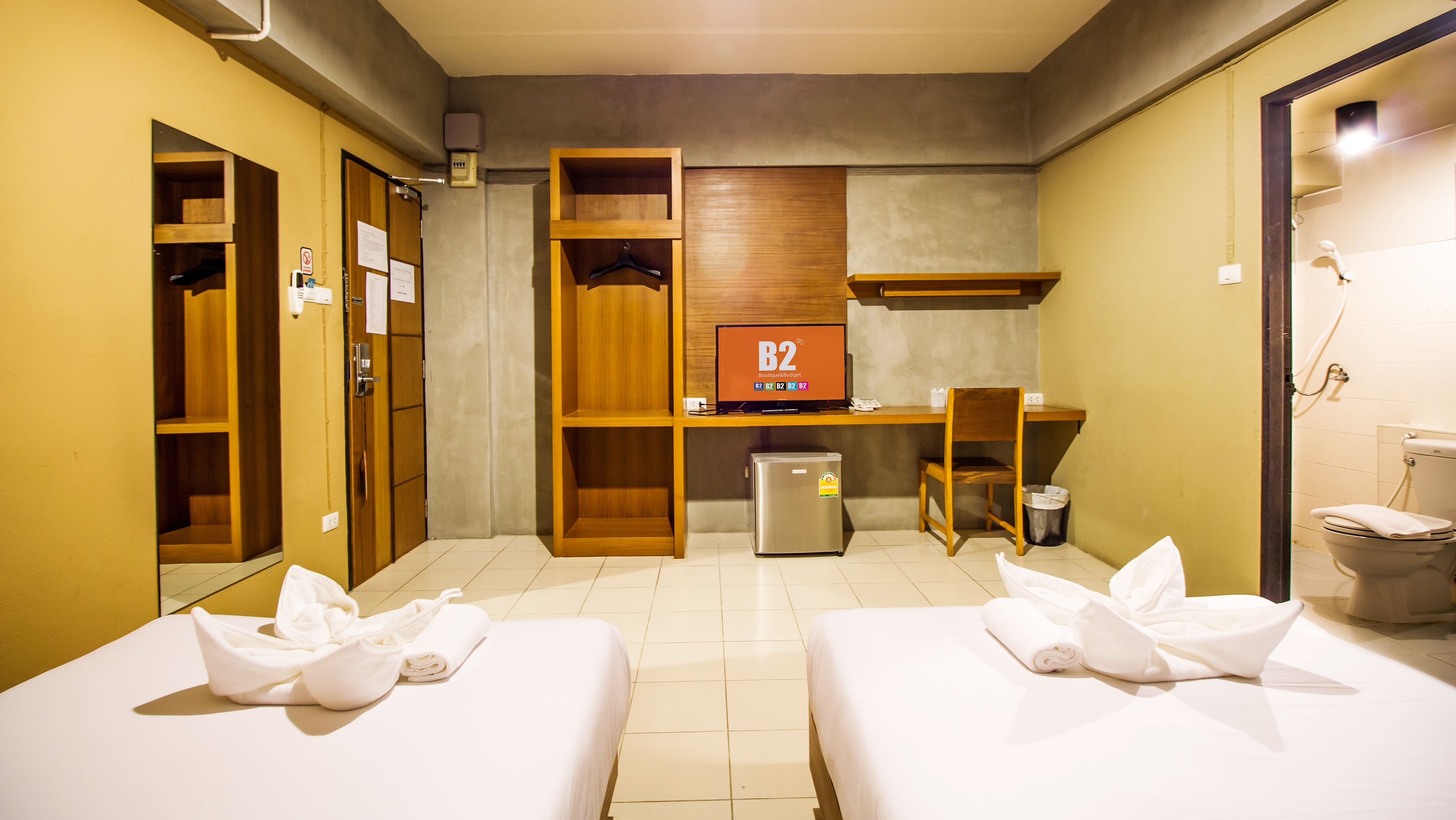 Ein Hotelzimmer mit zwei Betten, einem Schreibtisch, einem Stuhl, einer kleinen Kühlschrank, einem Spiegel und Holzschränken.