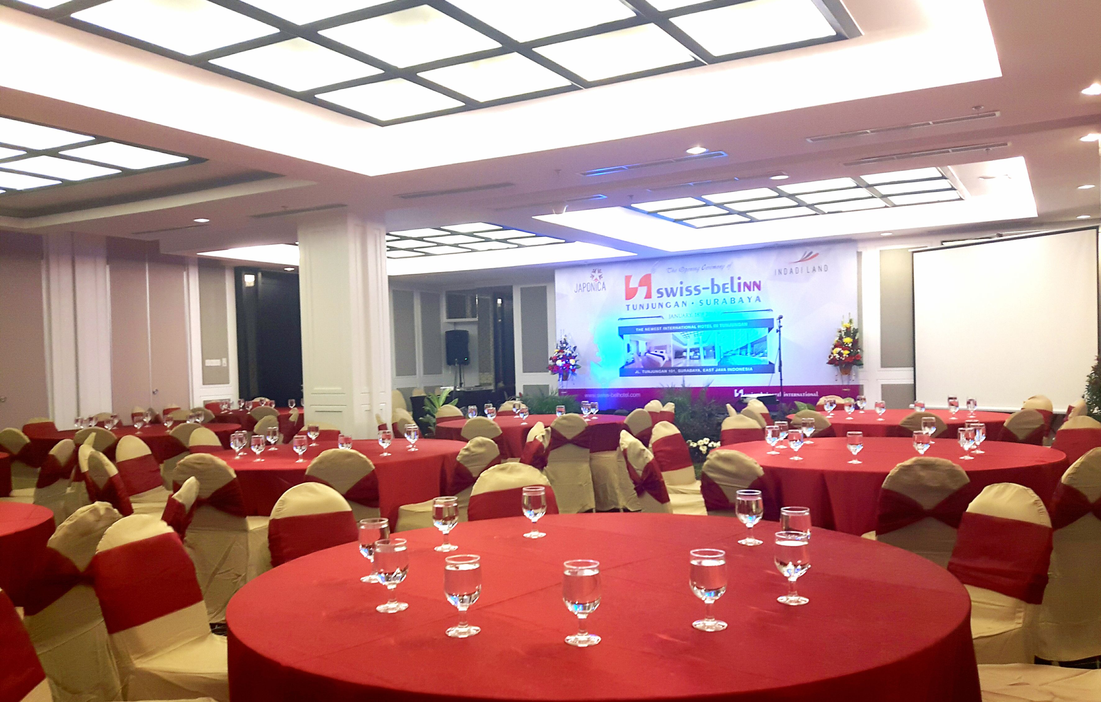 A conference room with round tables, red tablecloths, and chairs with red and white sashes. A stage with a banner is visible in the background.