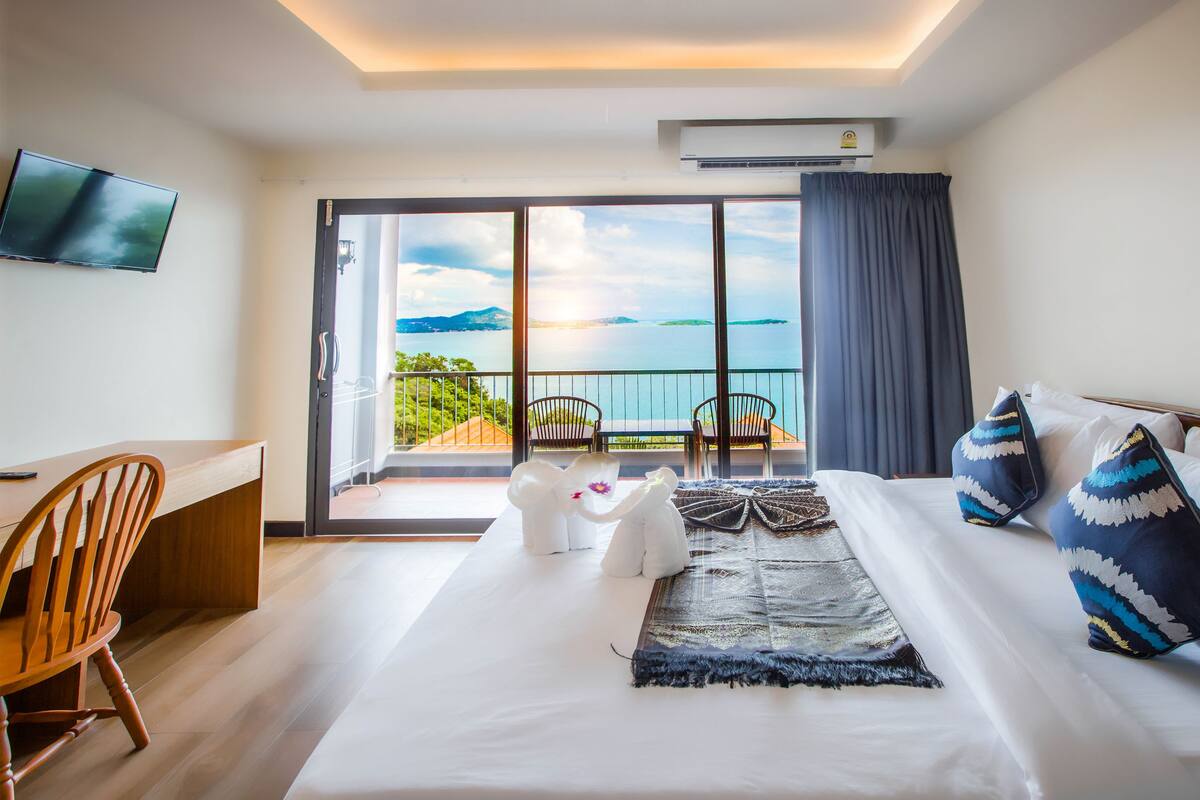 A hotel room with a bed, a desk, a chair, and a balcony with a view of the sea.