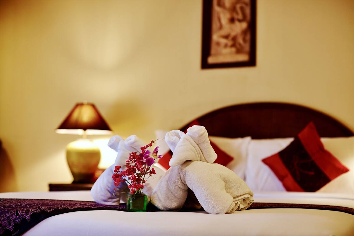 A neatly made bed with towels and a vase of flowers, a lamp, and a framed picture on the wall.