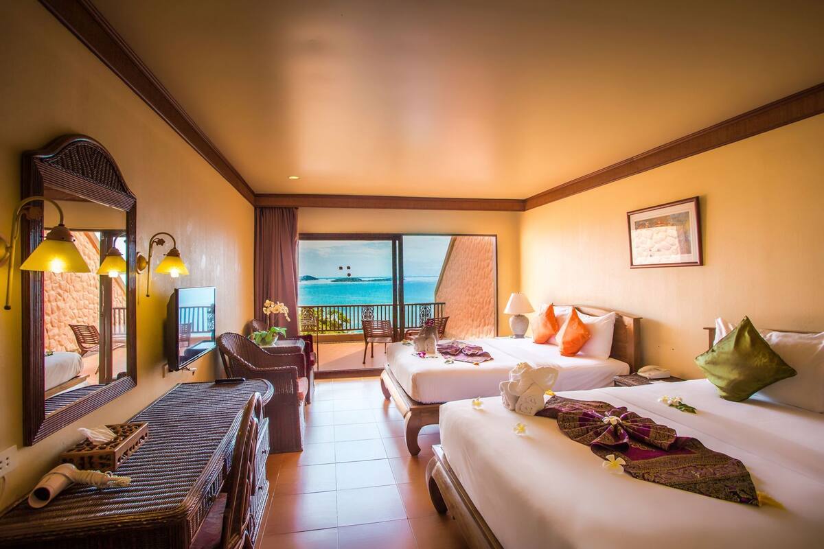 A hotel room with two beds, a balcony with a view of the sea, a desk with a chair, and a large mirror.