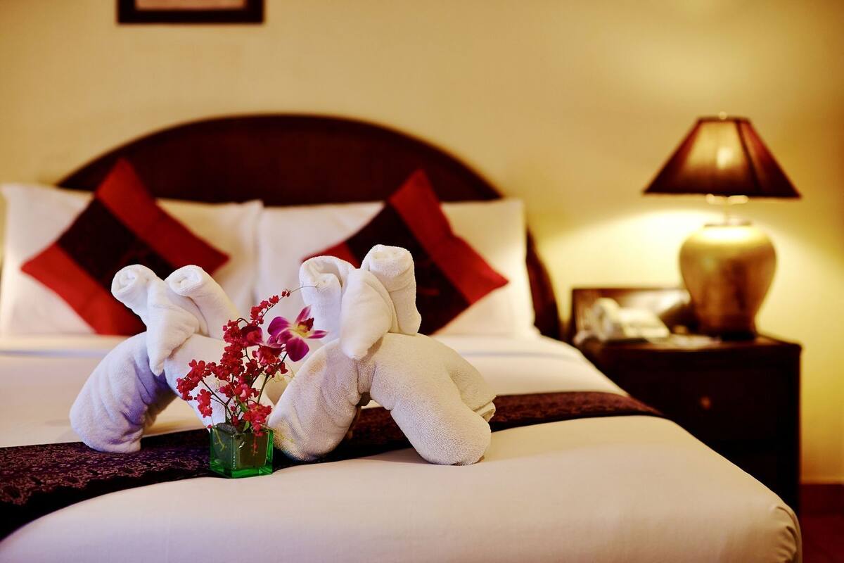 A neatly made bed with white linens, a red and black pillow, and a small vase with flowers.