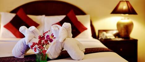 A neatly made bed with white and red pillows, a small vase with flowers, and a pair of white towels arranged in a bunny shape.