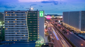 Un hotel de varios pisos con un letrero llamativo que dice 'HOTEL 101 MANILA', un edificio moderno con fachada verde y blanca, y vista a una calle concurrida con palmeras y una masa de agua al fondo.