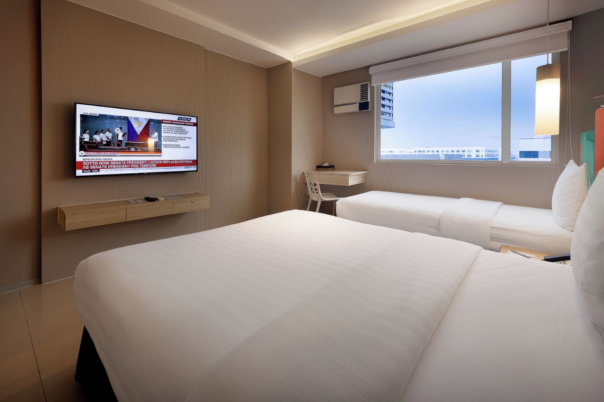 A hotel room with two beds, a TV mounted on the wall, a desk, and a window with a city view.