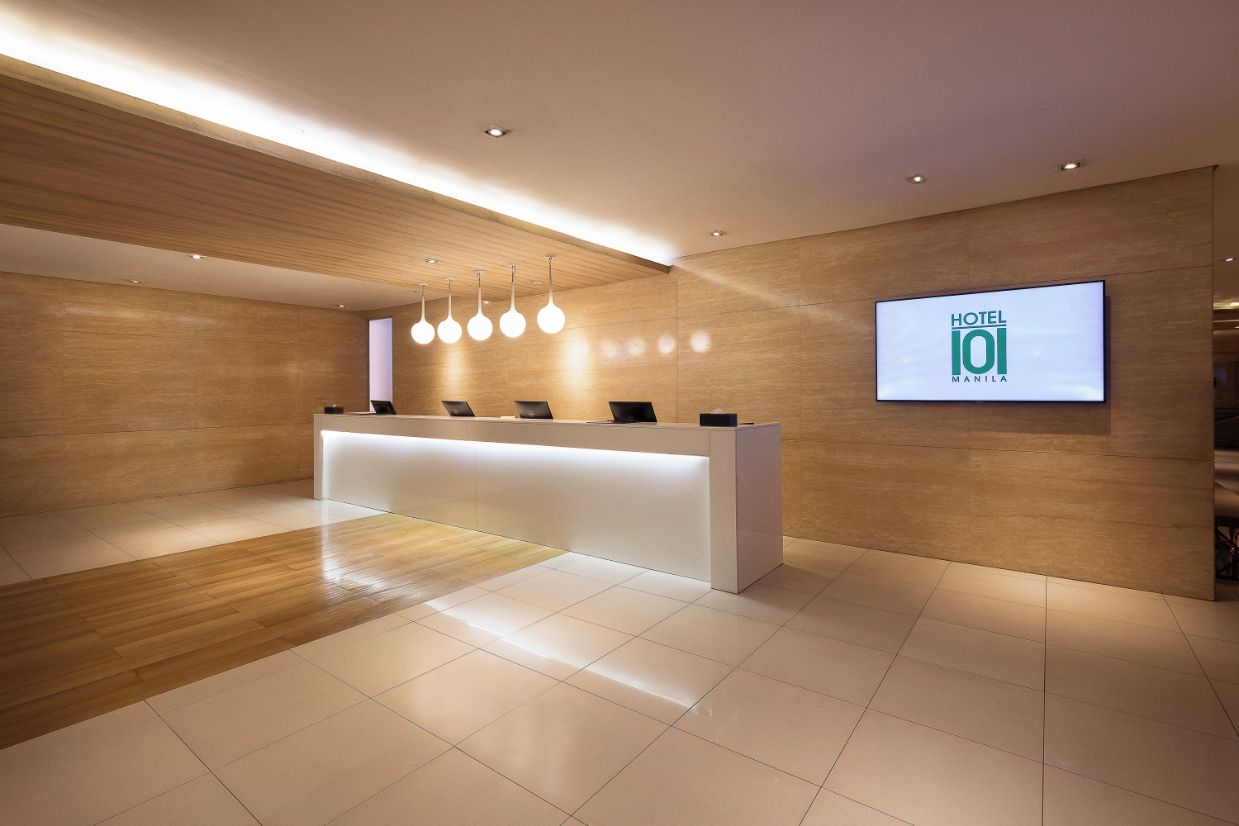 A modern hotel reception area with a wooden reception desk, a digital screen displaying the hotel name, and a clean, tiled floor.