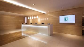 A modern hotel reception area with a wooden reception desk, a digital screen displaying the hotel name, and a clean, tiled floor.