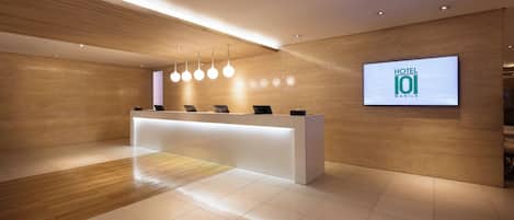 A modern hotel reception area with a wooden reception desk, a digital screen displaying the hotel name, and a clean, tiled floor.
