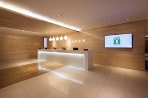 Ein moderner Hotel-Empfangsbereich mit einem Holzbüro, einem digitalen Bildschirm mit dem Hotelnamen und einem sauberen, gefliesten Boden.