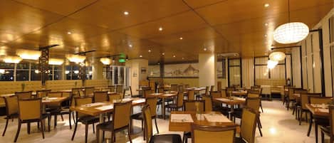 A spacious restaurant with multiple dining tables and chairs, a high ceiling with recessed lighting, and a large mural on the back wall.
