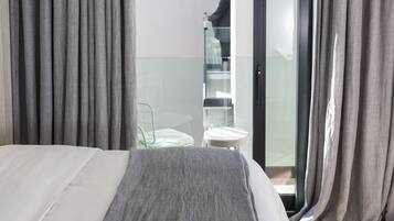 A hotel room with a bed, grey curtains, a glass door leading to a balcony, and a person standing by the door.
