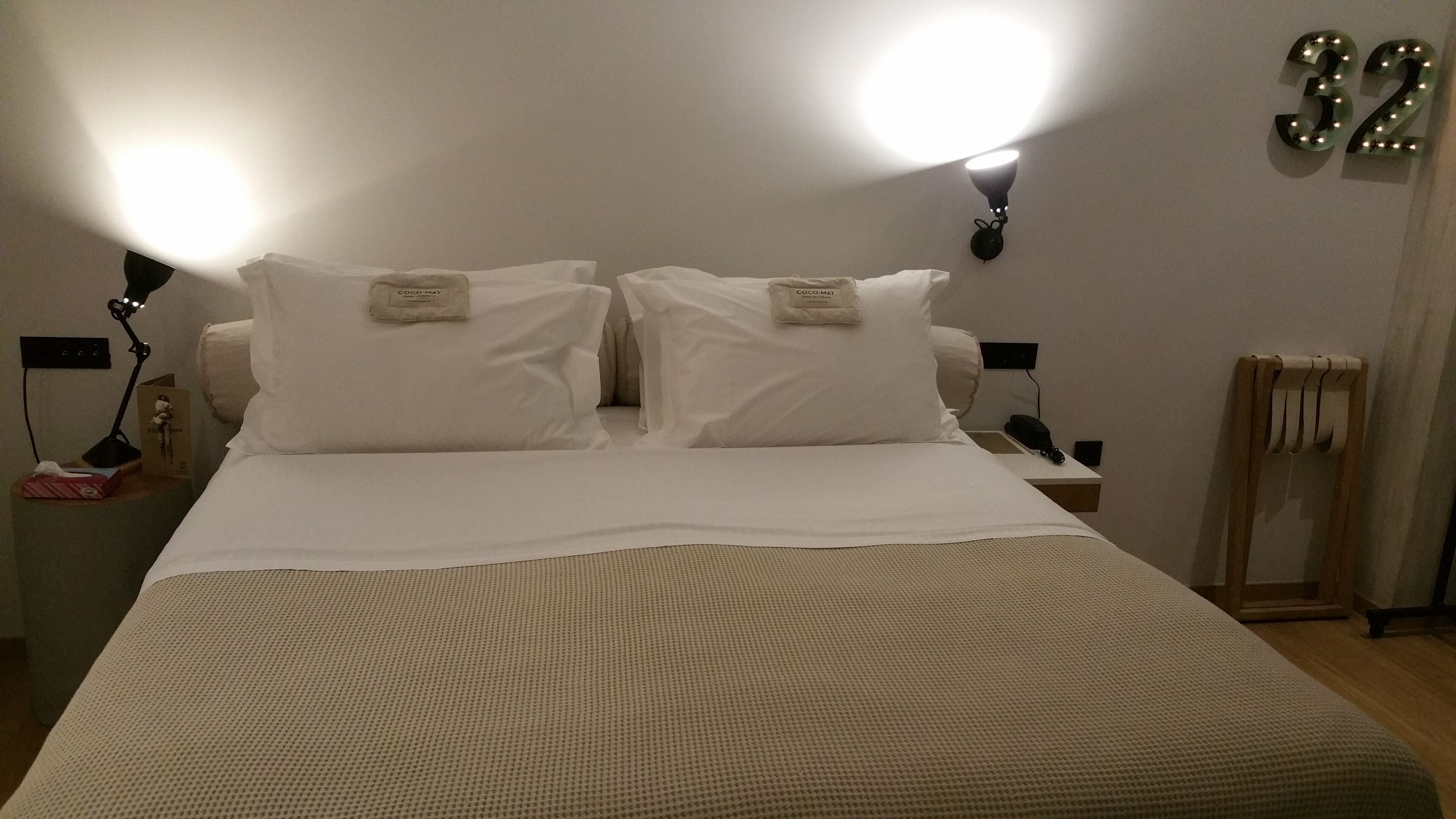 Una cama bien hecha con sábanas blancas, dos lámparas fijadas a la pared y un cartel con el número 32.