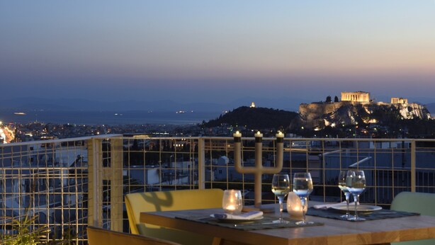 A table with wine glasses and a menorah, overlooking a cityscape and ancient ruins at dusk.