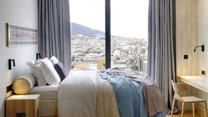 A modern hotel room with a large bed, a desk, and a view of a cityscape through floor-to-ceiling windows.