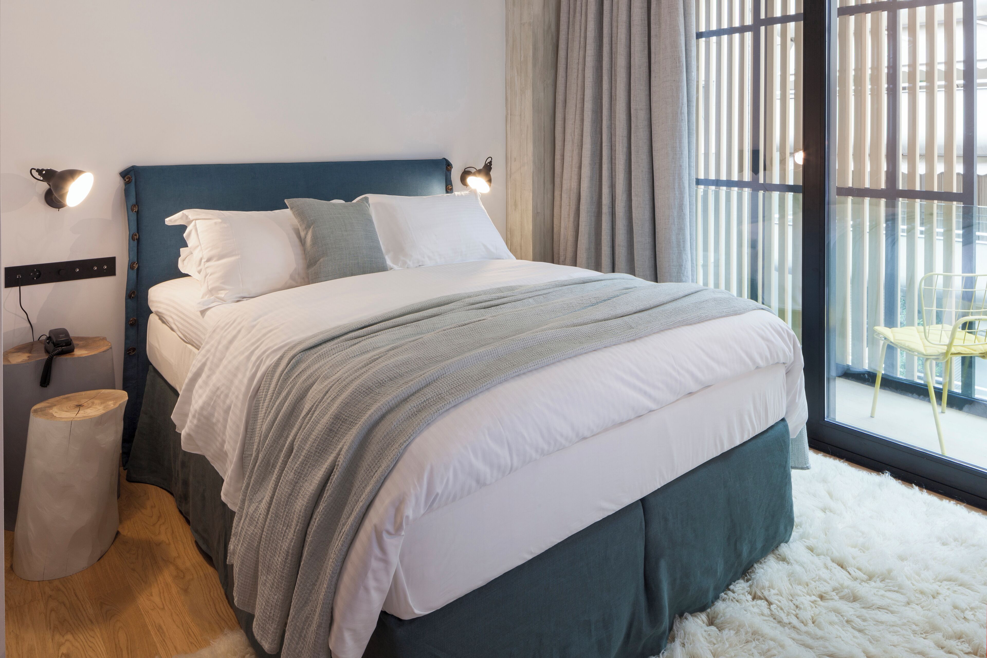 A modern bedroom with a large bed, a nightstand, a lamp, and a view through a window.