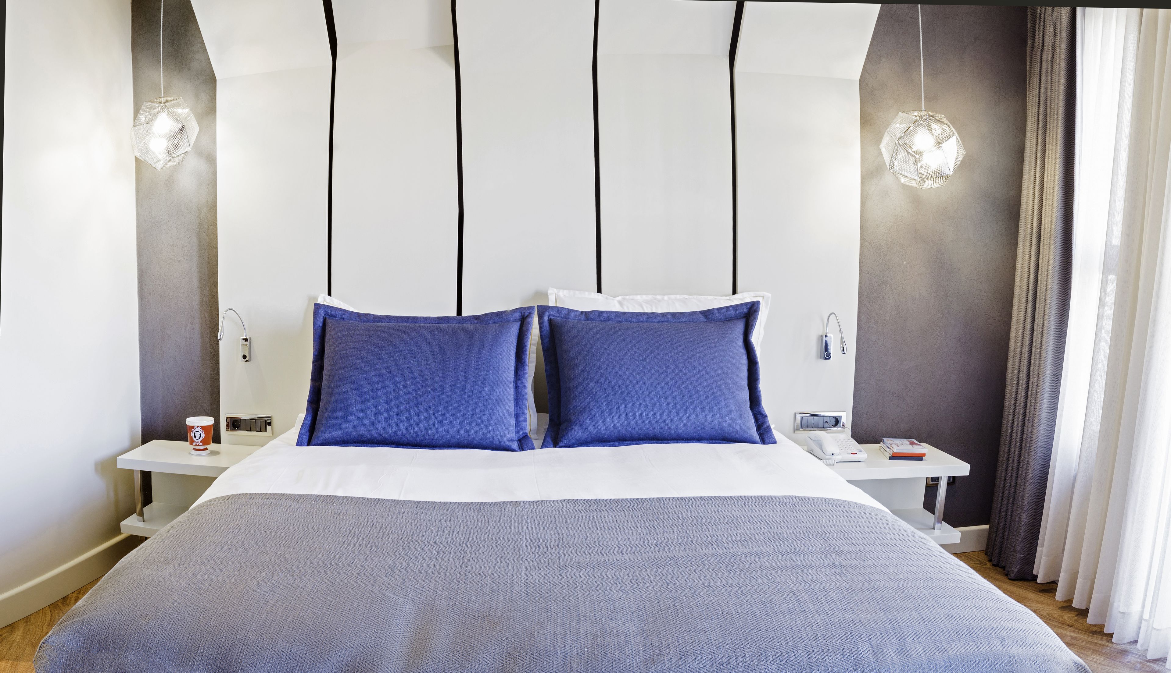 Habitació d'hotel moderna amb un llit gran, dos coixins blaus, una manta blanca, una taula de nit amb una làmpada i una estació de nit amb un telèfon i llibres.
