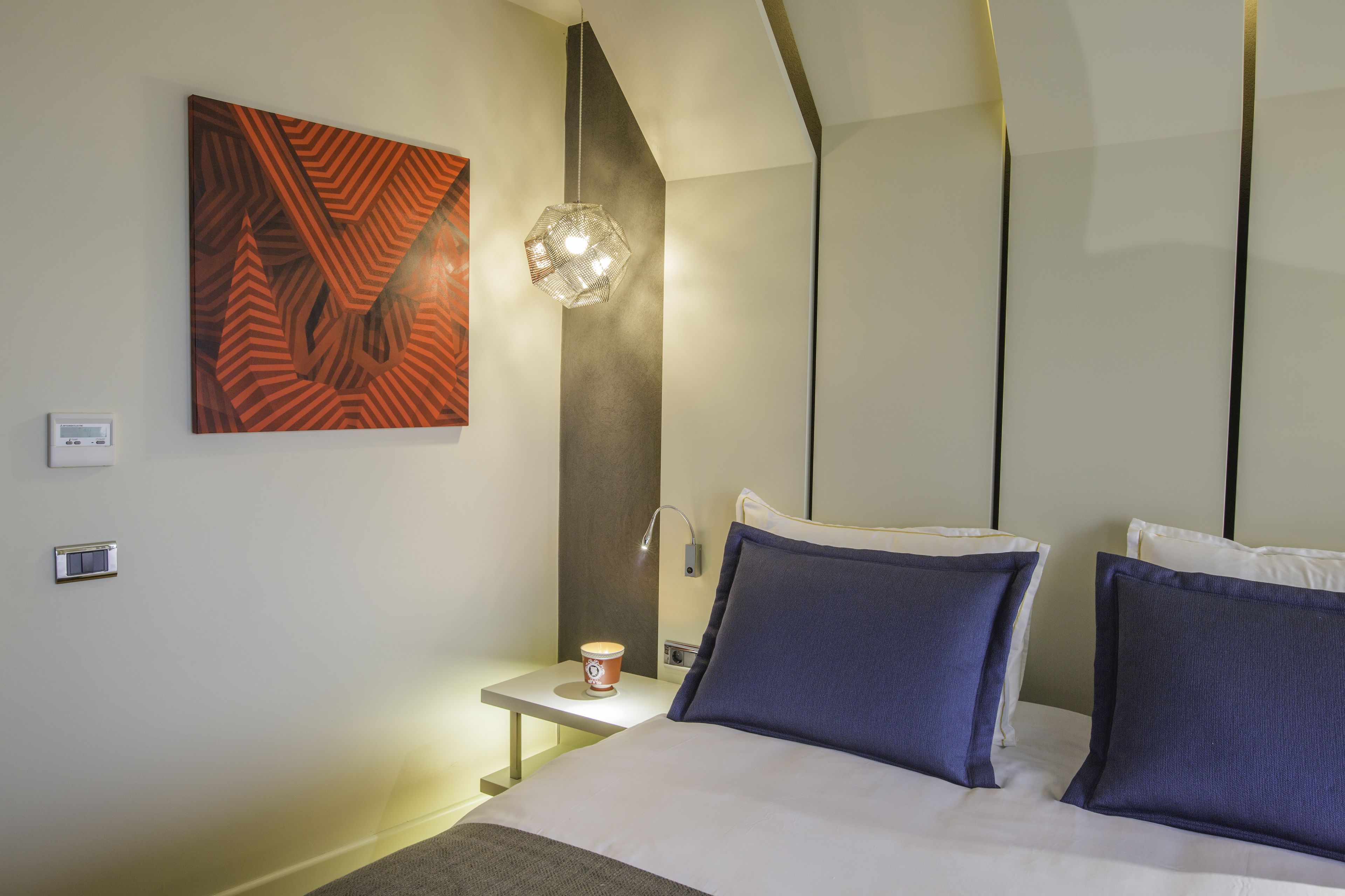 A modern hotel room with a large bed, two pillows, a bedside table with a cup, a wall-mounted artwork, and a unique light fixture.