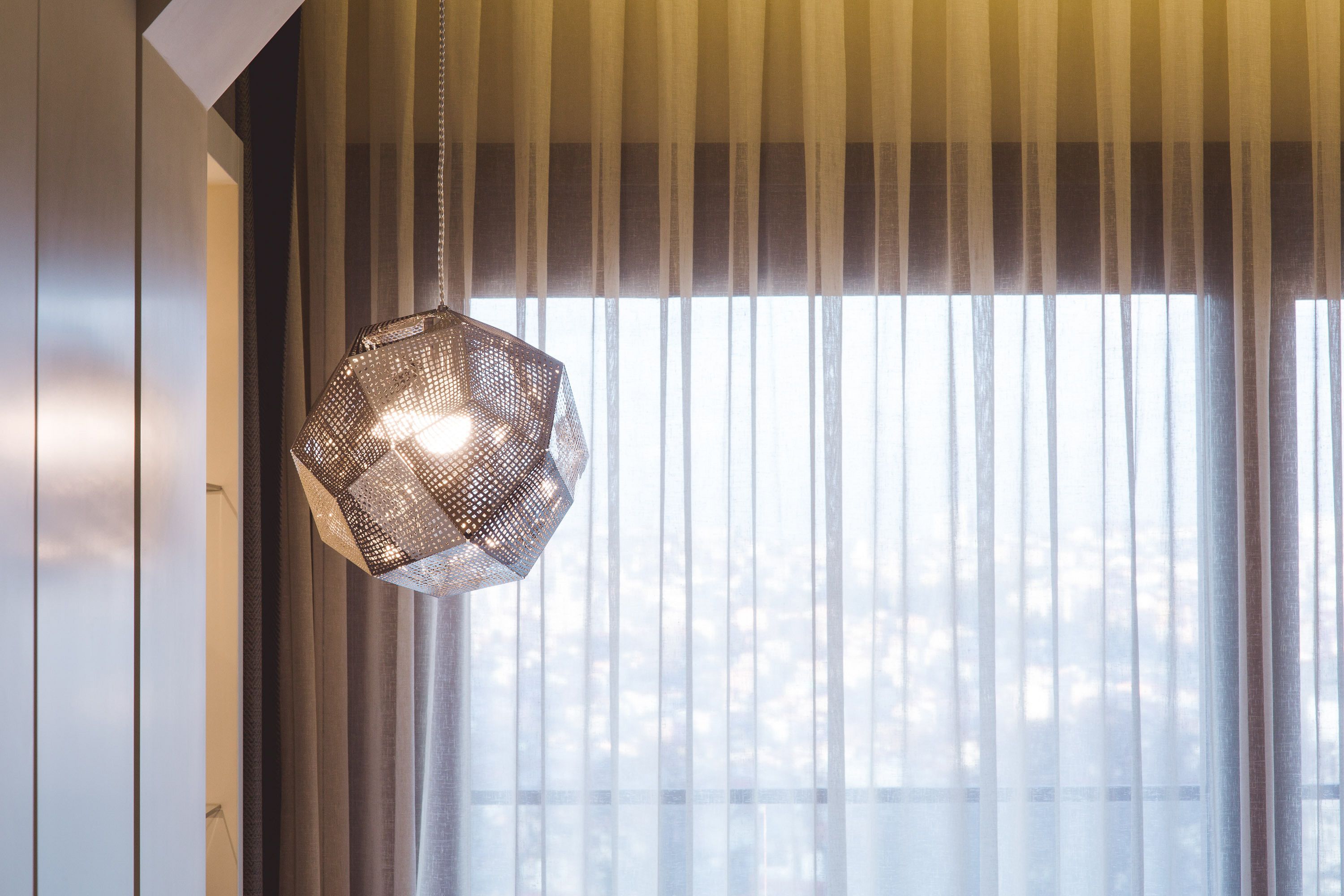 A hanging light fixture with a geometric design, a window with vertical blinds, and a view of trees outside.