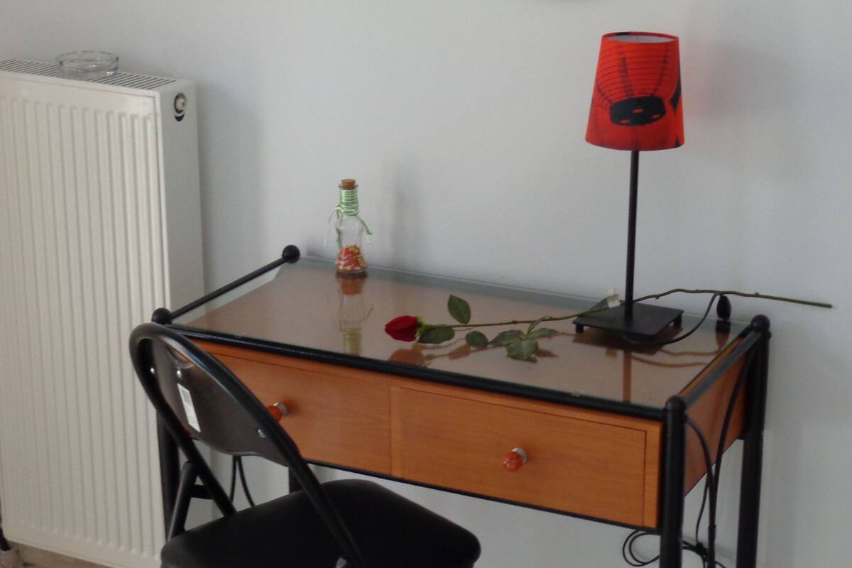 Un pequeño escritorio de vidrio transparente, una silla negra, un radiador, una lámpara roja y una rosa sobre el escritorio.