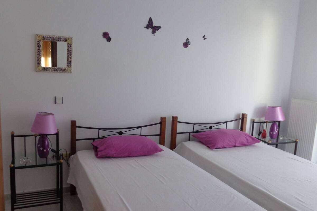Una habitación con dos camas, cada una con un colchón blanco y almohadas moradas, y una mesita con una lámpara morada. Hay un espejo en la pared y decoración con mariposas.