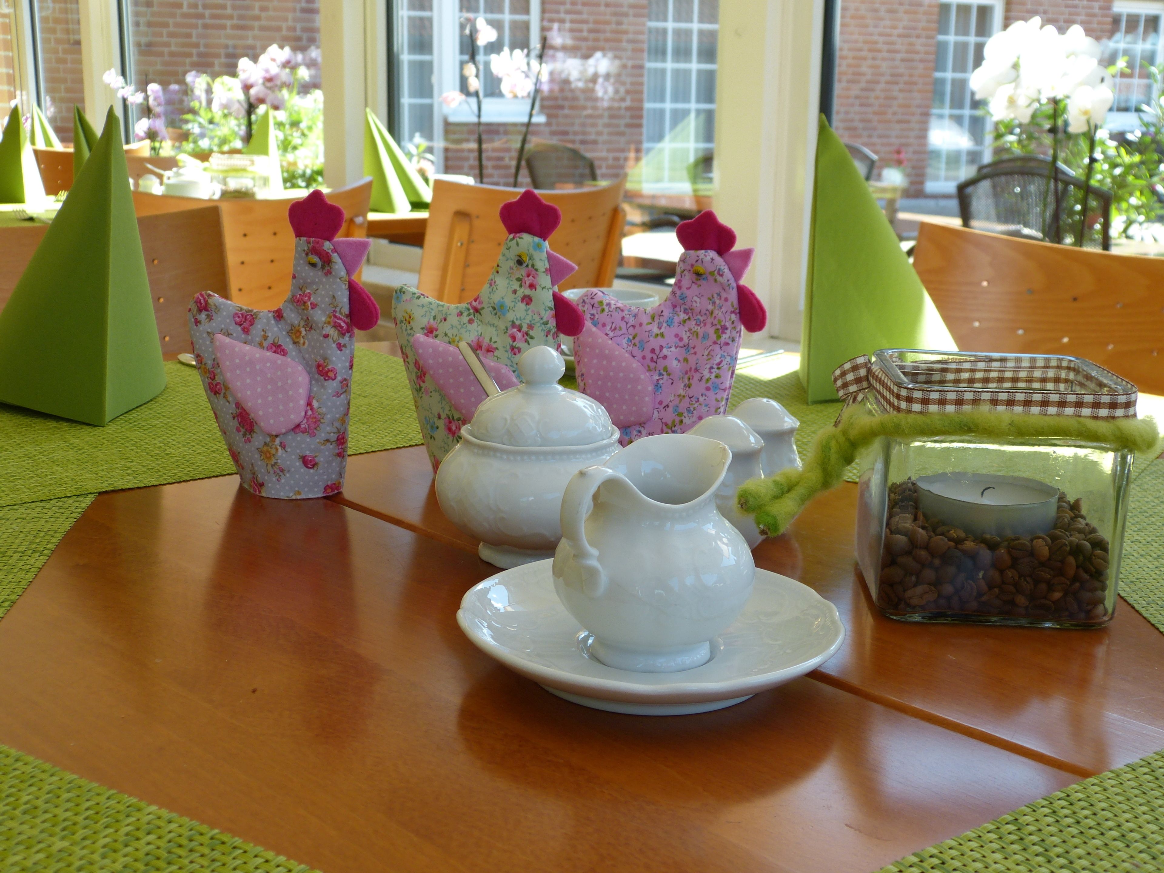 A table set with white ceramic teapot, jug, and cups, surrounded by floral-patterned napkins and a glass jar with coffee beans.