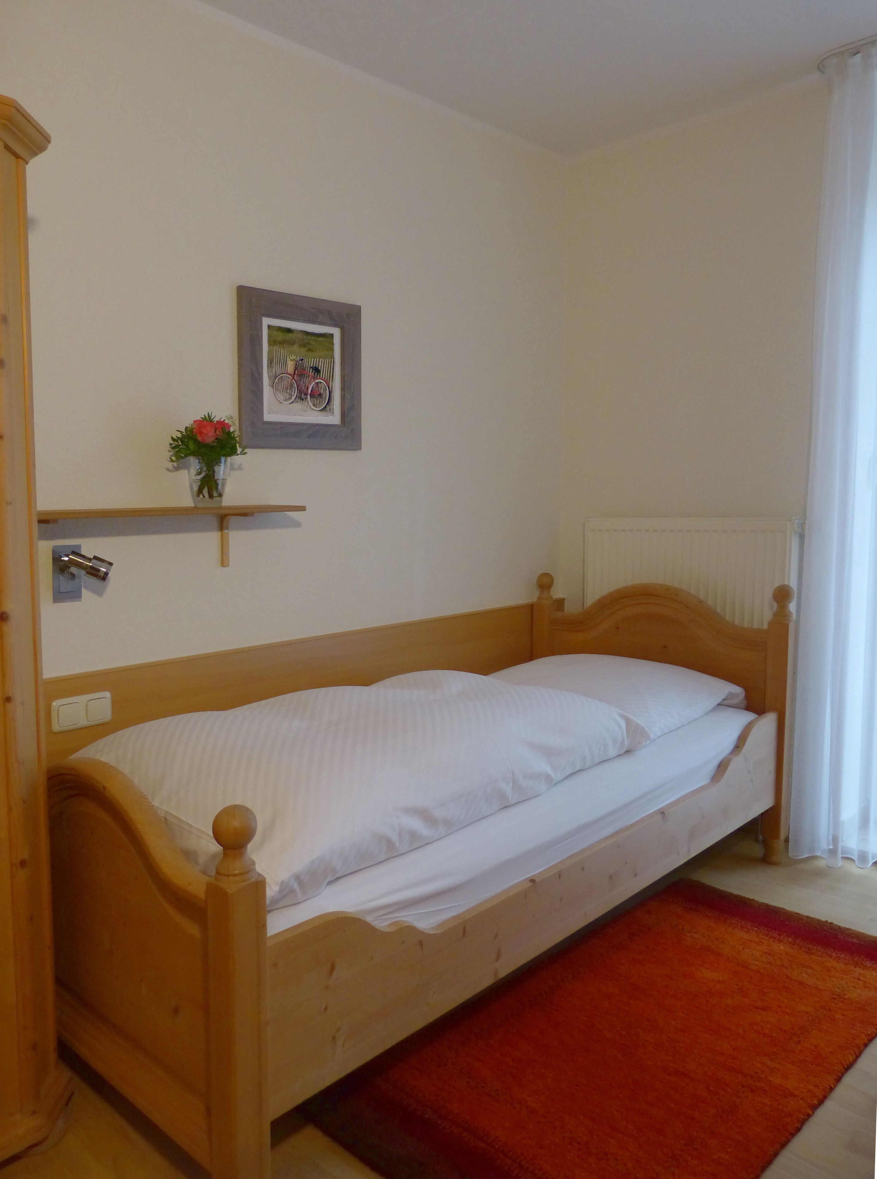 A single wooden bed with white bedding, a red rug, a small shelf with a flower, and a framed picture on the wall.