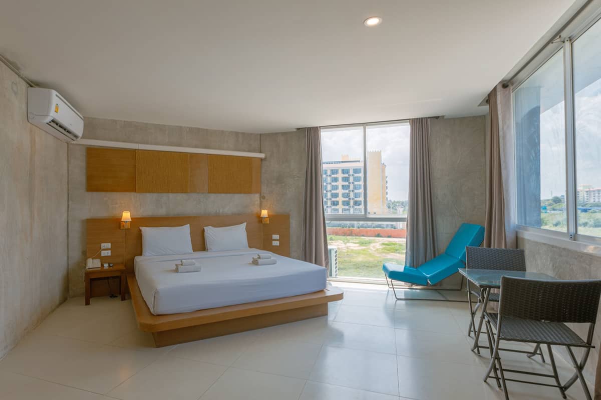 A modern hotel room with a large bed, a desk with a chair, and a view of the city through large windows.