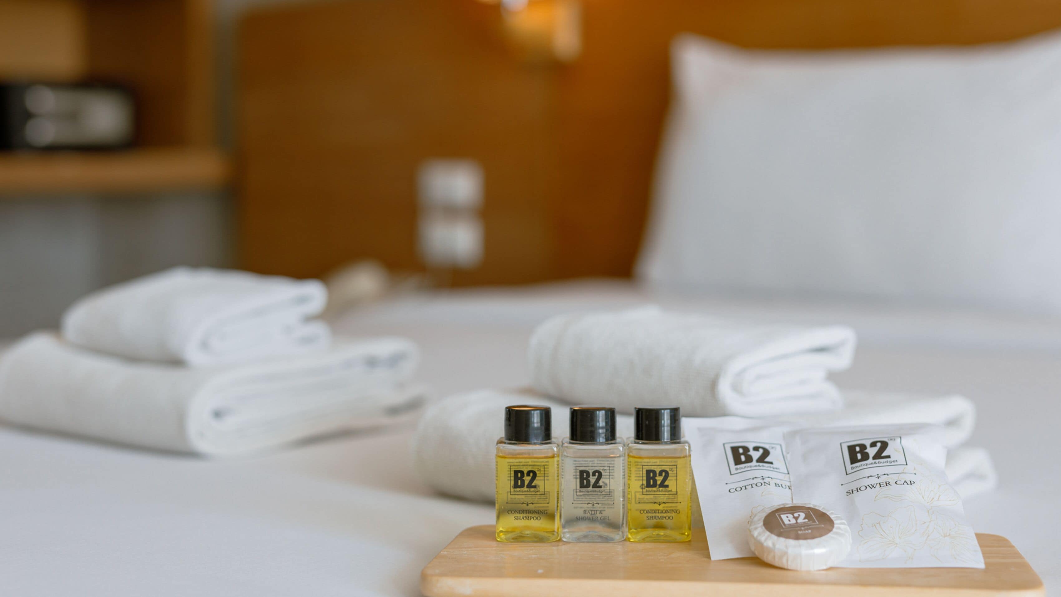 A neatly made bed with folded white towels, a wooden tray with three bottles of B2 conditioning shampoo, a bottle of B2 shower gel, and a B2 cotton bud.