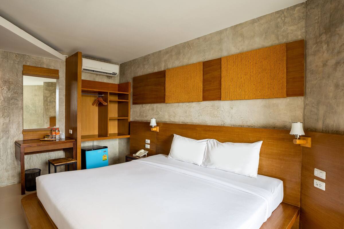 A hotel room with a large bed, a wooden wardrobe, a desk with a mirror, and a small blue minibar.