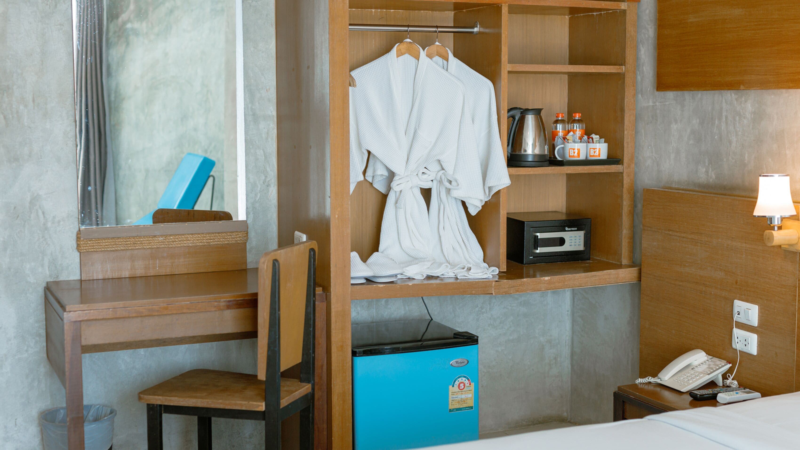 Une chambre d’hôtel avec un bureau en bois, une chaise, un mini-bar bleu, un peignoir suspendu à un porte-manteau, une machine à café et un téléphone sur la table de chevet.