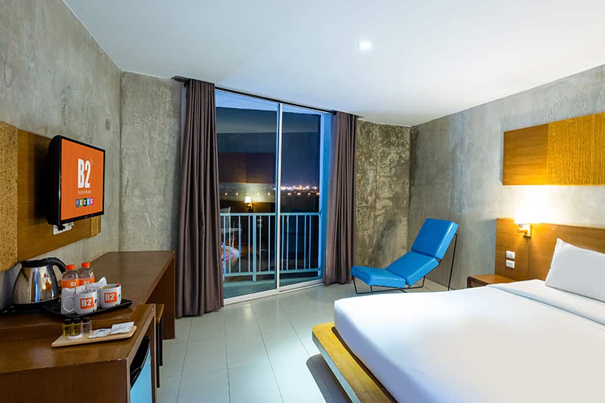 A hotel room with a large bed, a desk with a TV, a blue chair, and a balcony with a view of the city at night.