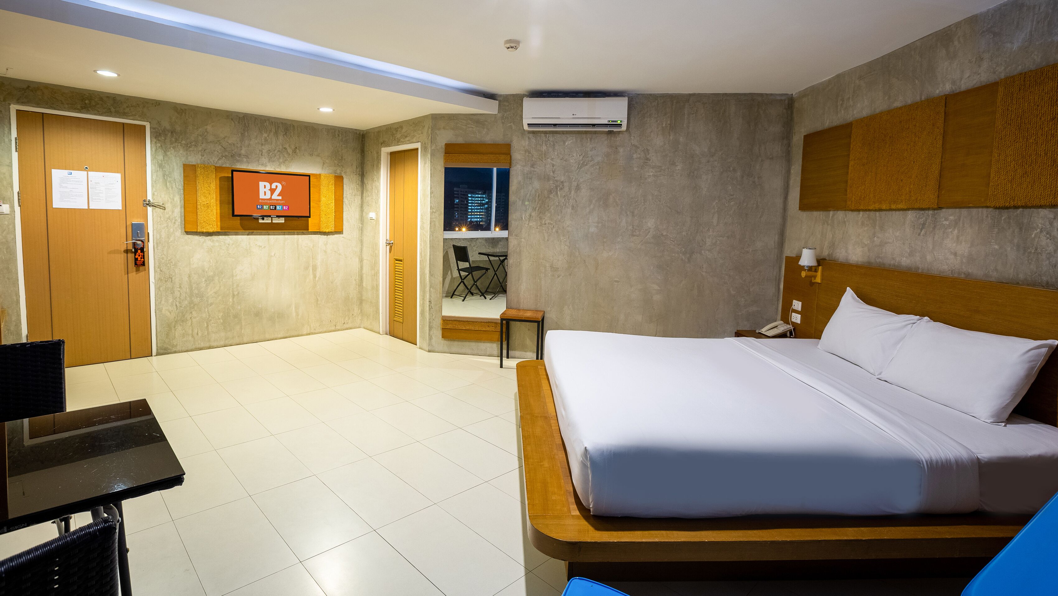 Habitación de hotel con cama, escritorio, silla, mesita, mesita de noche, lámpara y vista a la ciudad de noche.