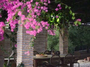Une terrasse extérieure avec une table en briques et des chaises en osier, entourée d’une végétation luxuriante et de plantes grimpantes en fleurs.