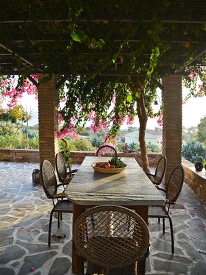 Une terrasse extérieure avec un sol en pierre, une table en bois, des chaises en osier et une vue sur un jardin planté de fleurs.