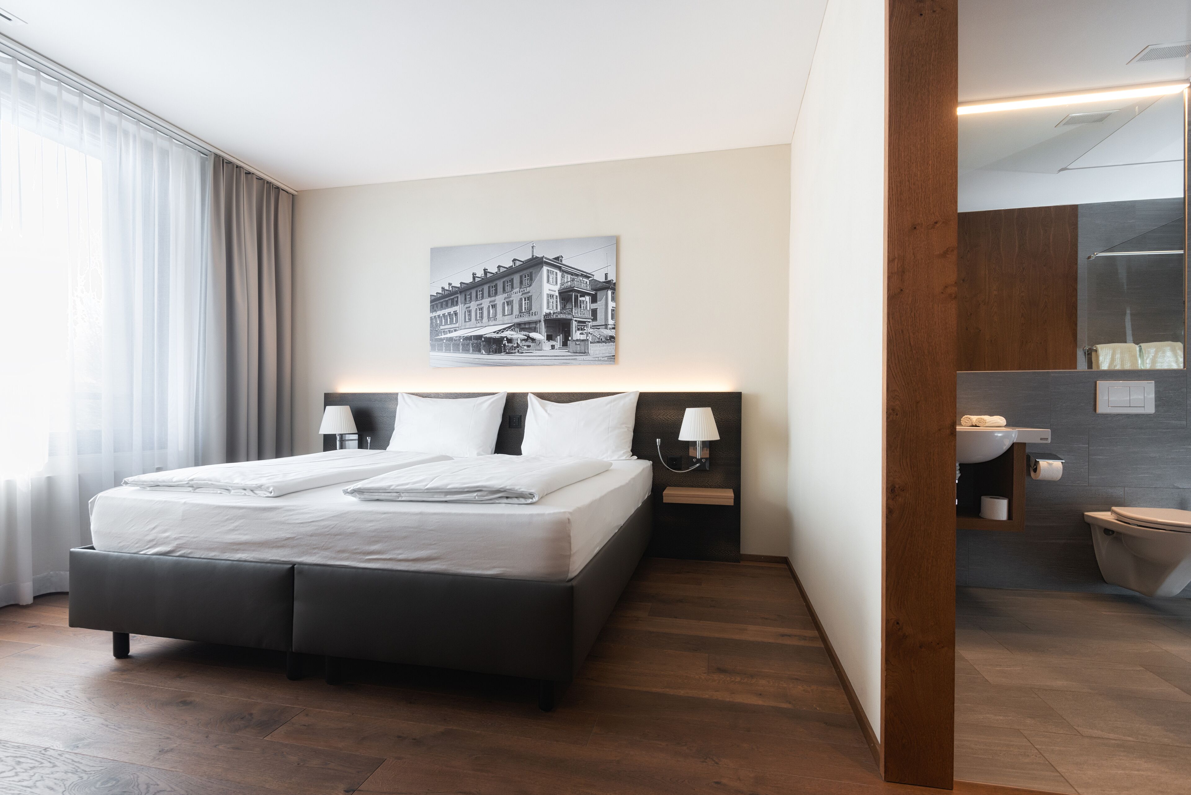 A modern hotel room with a large bed, a nightstand with a lamp, a bathroom with a mirror, and a wall-mounted picture.