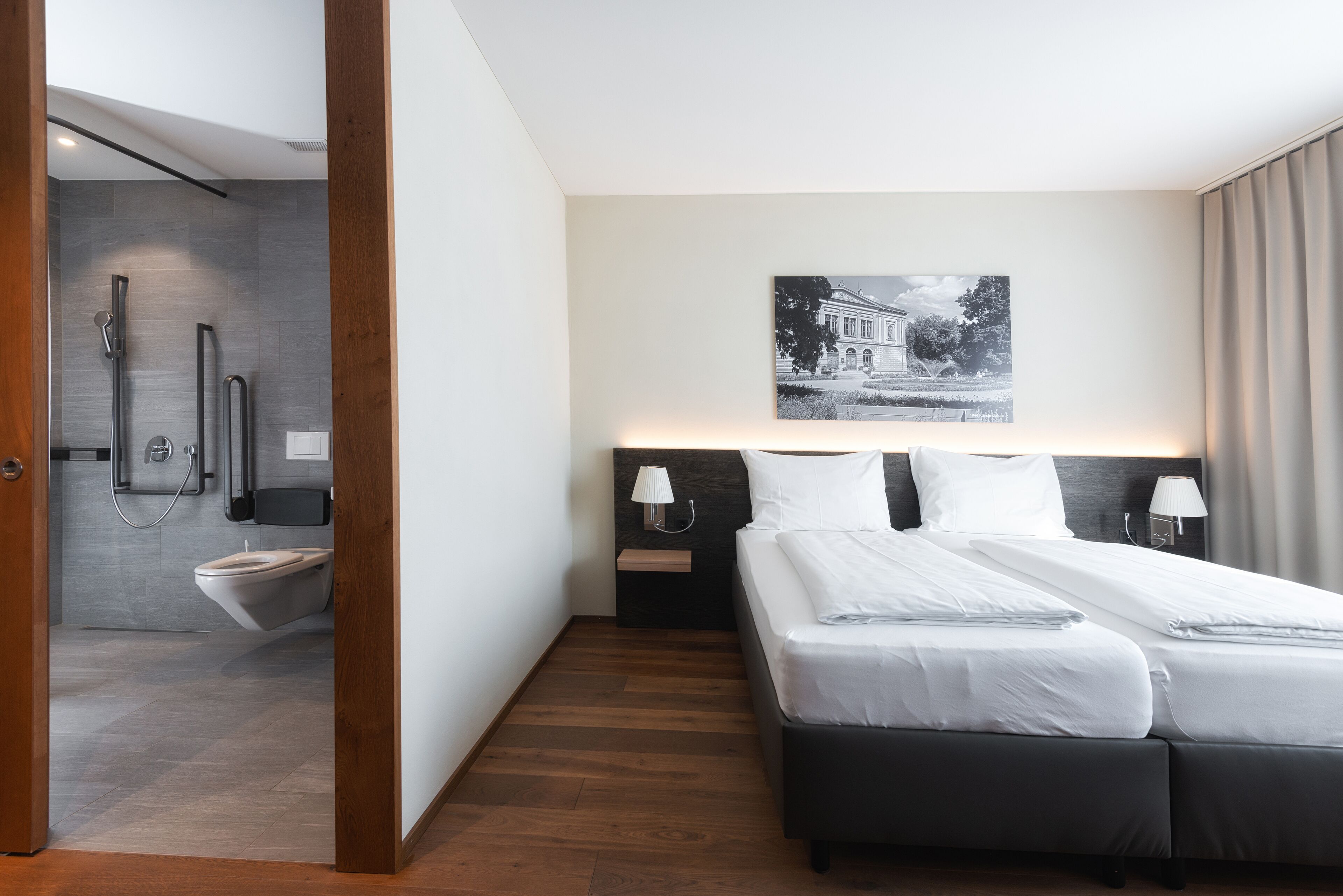 A modern hotel room with a large bed, a nightstand, a wall-mounted lamp, and a picture of a building.