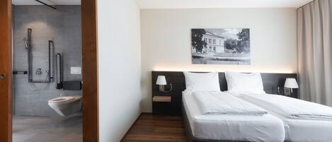 A modern hotel room with a large bed, a nightstand, a wall-mounted lamp, and a picture of a building.